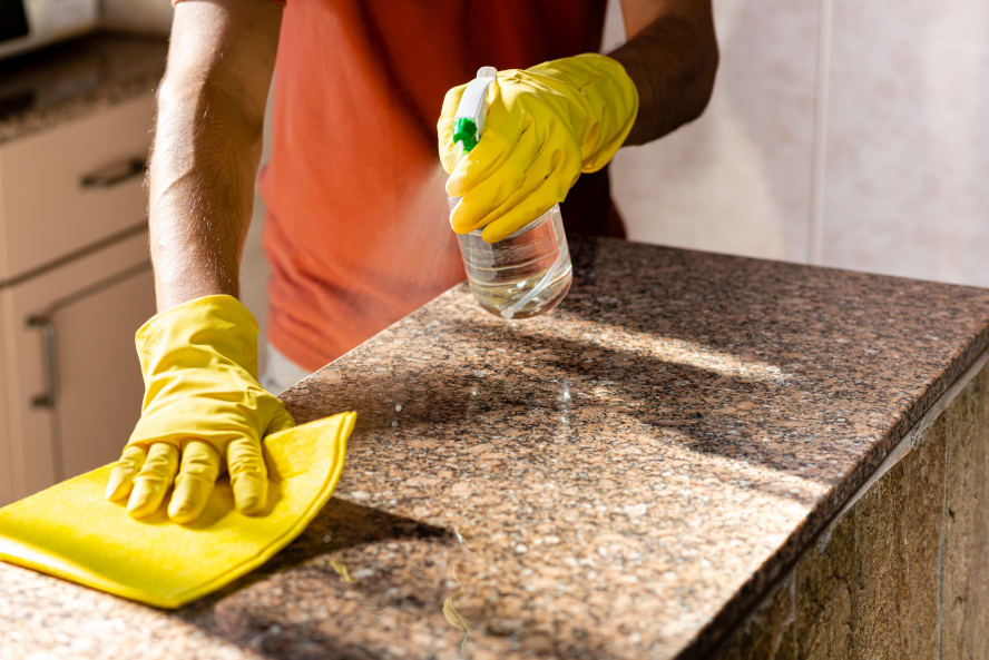 person wearing orange t-shirt and yellow rubber gloves holding spray bottle cleaning granite countertop