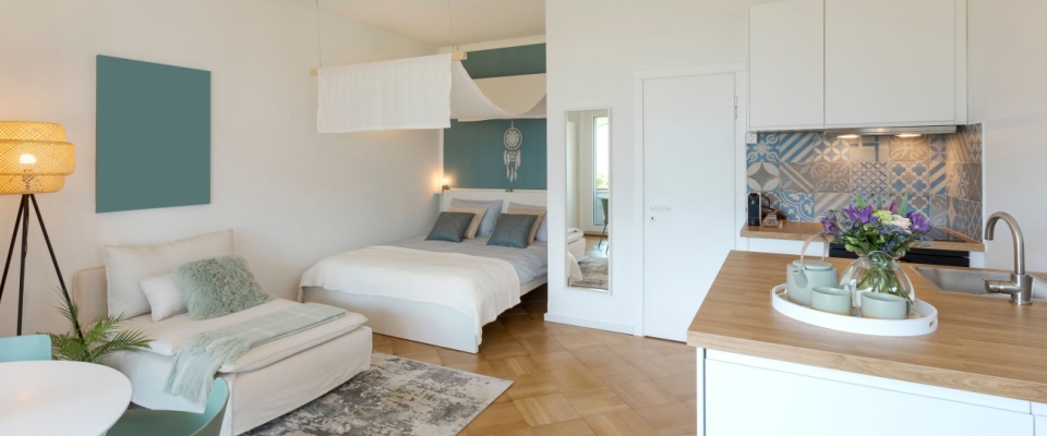 General view of a stylish studio apartment. There is a small kitchen with a wooden counter, a double bed and a single bed, and a table with chairs.