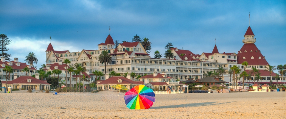 A rainbow-colored umbrella sits on the sandy Coronado Beach, a top San Diego destination for renters, with the grand Hotel del Coronado visible under a pleasant sky.