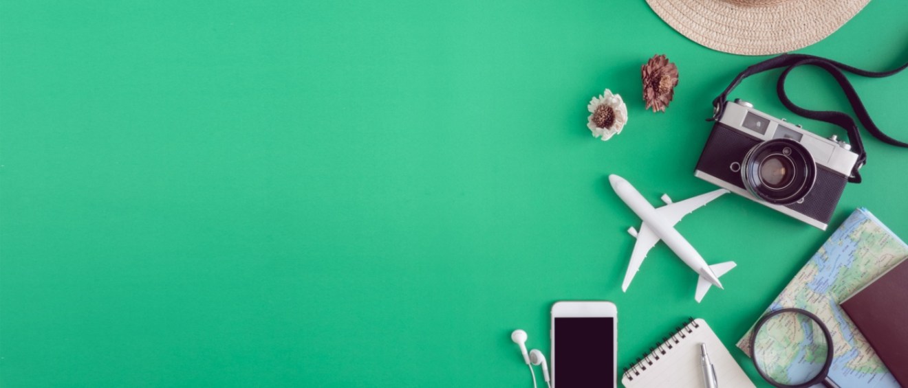 Pre-travel items on a green background: a straw hat, vintage camera, pine cones, toy airplane, smartphone, notebook, map, and magnifying glass, suggesting adventure and planning