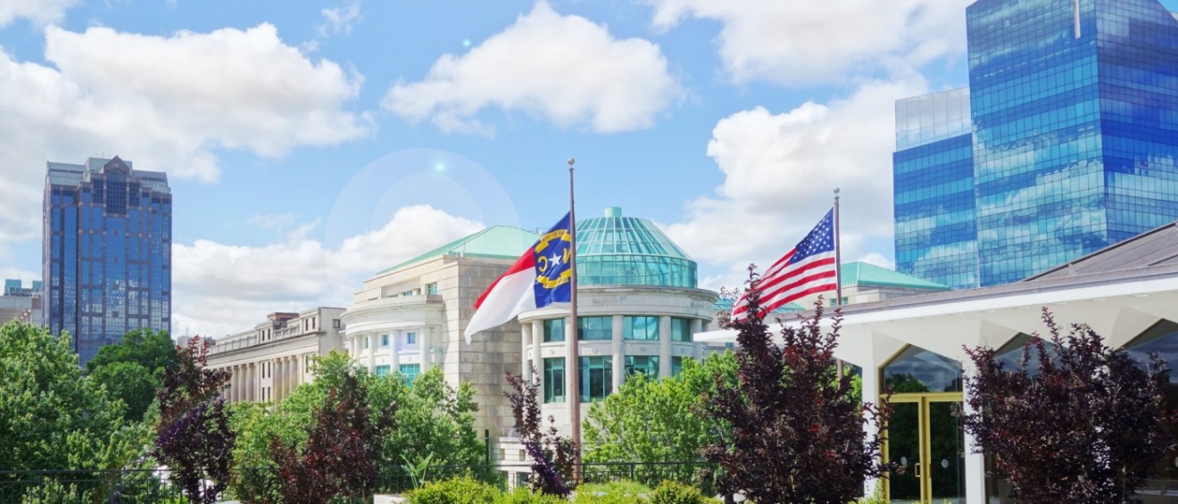 Downtown Raleigh, North Carolina, featuring modern buildings, the North Carolina Museum of Natural Sciences, and state and American flags against a bright, partly cloudy sky.