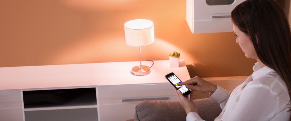 A woman uses her mobile phone to control the lighting in her rental apartment, which includes a table lamp on a modern white cabinet.