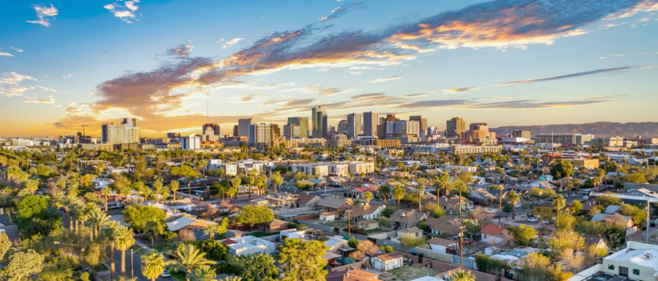 Aerial view of Phoenix, AZ, showcasing a vibrant city skyline with high-rise buildings set against a backdrop of a colorful sunset, residential neighborhoods with lush green trees, and palm trees lining the streets in the foreground.