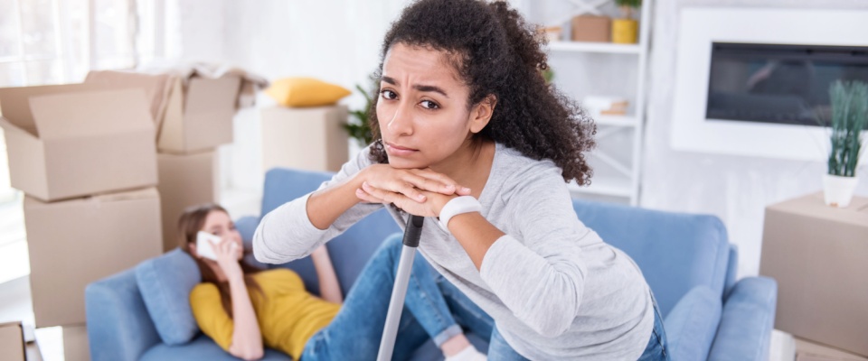Girl cleaning the floor in the new apartment while her roommate lying on the sofa and doing nothing but talking on the phone.