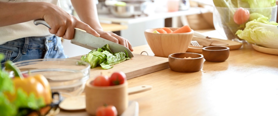 A close-up image of a woman cutting lettuces on a cutting board at a wooden kitchen tabletop, cooking in the kitchen.