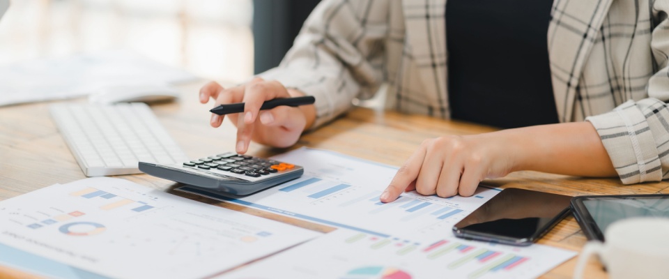 Businessperson calculating budget with a calculator and analyzing financial charts and graphs on documents at a wooden desk.