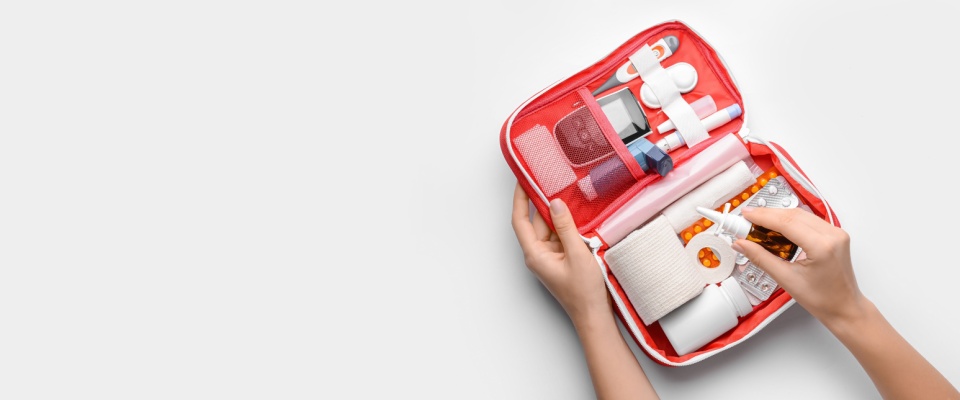 A person opens a well-organized red first aid kit, revealing various medical supplies including bandages, gauze, pills, and a nasal spray, all neatly arranged in compartments.