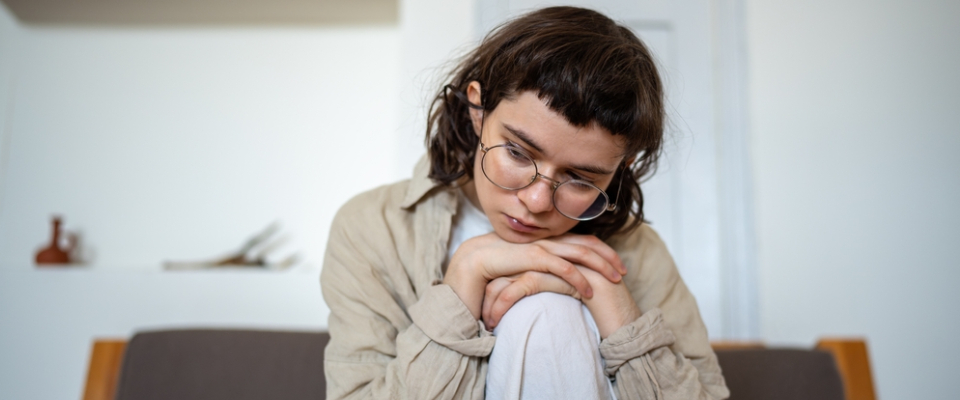 A young woman sitting on a couch in her new apartment, resting her chin on her knees and looking down thoughtfully, reflecting the emotional ups and downs of moving into her first home.