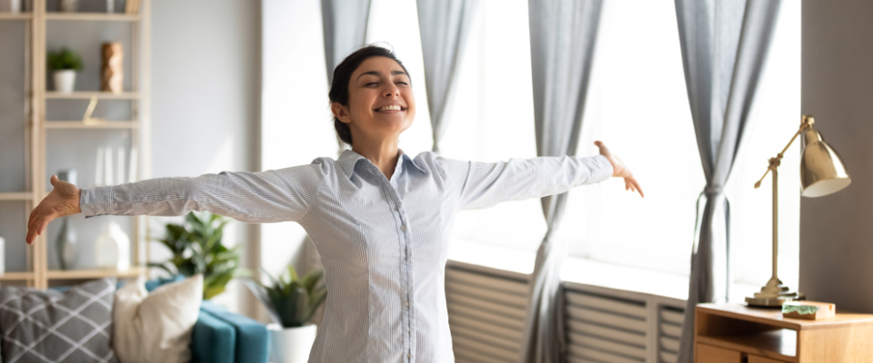 A young renter stands in her sunlit apartment with arms wide open and a big smile, feeling happy and free after moving into her first apartment.