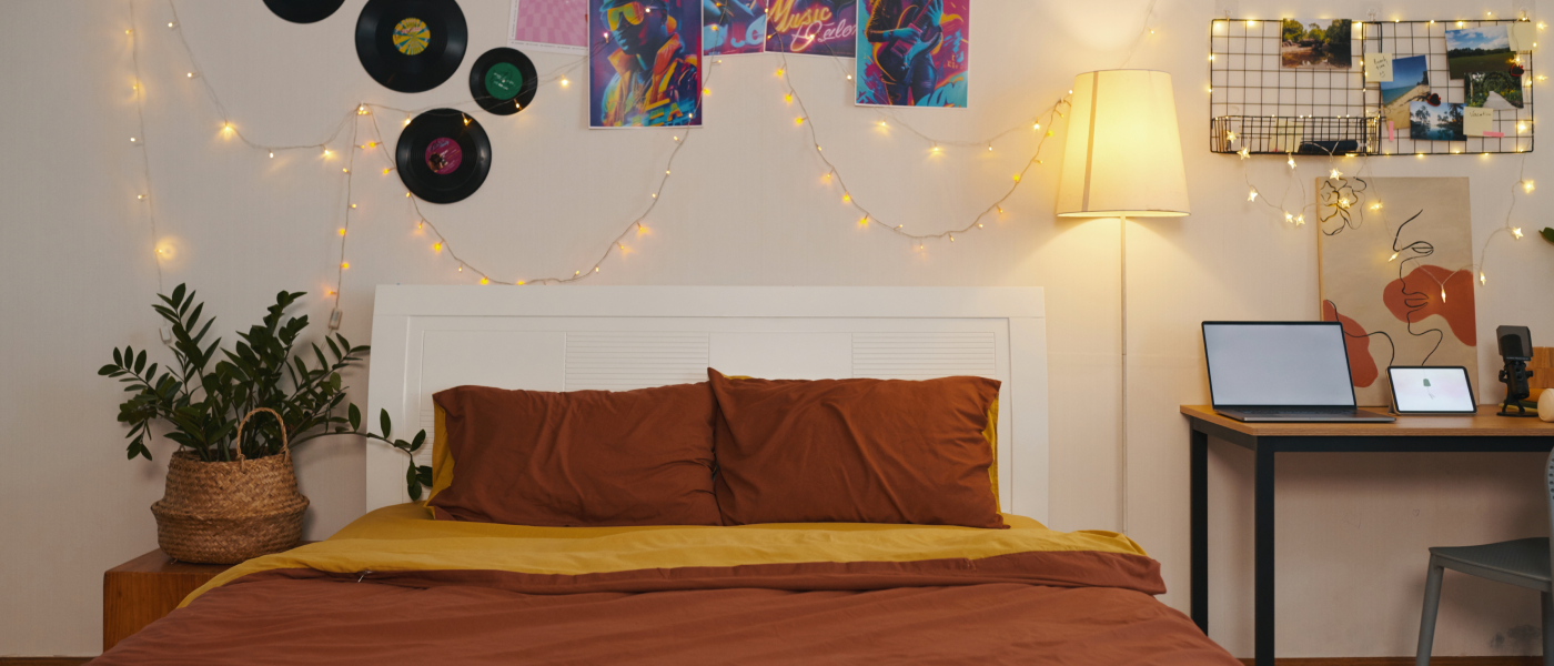 A cozy bedroom setup in a first apartment featuring a bed with brown and mustard bedding, string lights, vinyl records, and colorful posters on the wall. A desk with a laptop, tablet, and lamp sits to the right, creating a warm and inviting atmosphere.