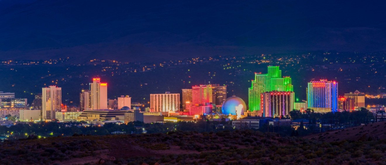 Nighttime view of Reno, Nevada's illuminated skyline with colorful buildings and surrounding desert landscape.