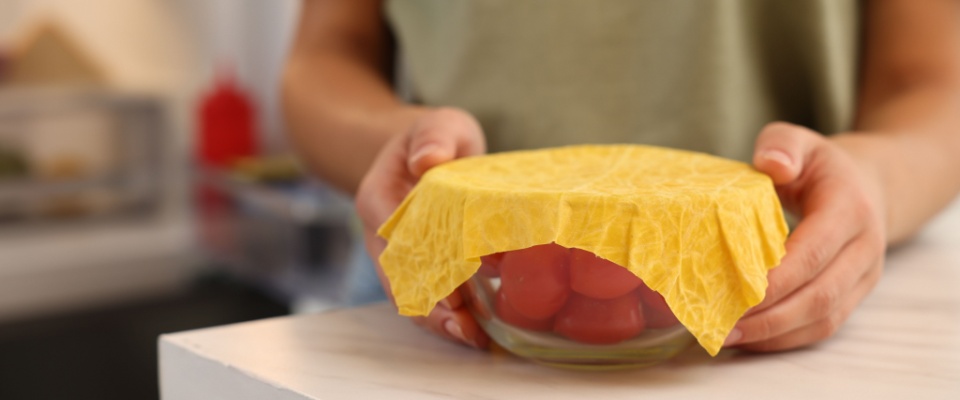 Woman holding bowl of fresh tomatoes covered with beeswax food wrap at table in kitchen.