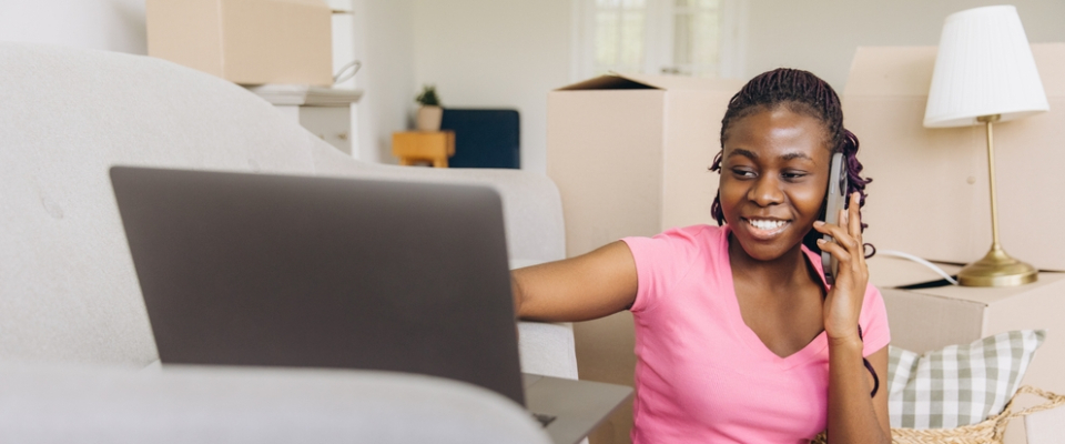 A young woman sitting on the floor surrounded by moving boxes, smiling while talking on the phone and looking at her laptop—possibly calling the landlord or handling apartment arrangements.