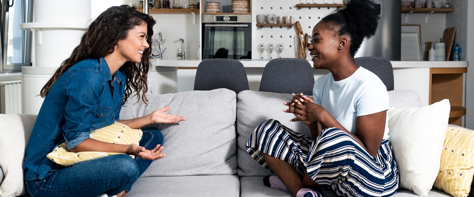 Two young women sitting on a couch in a modern apartment, smiling and having a friendly conversation, suggesting a positive roommate relationship.