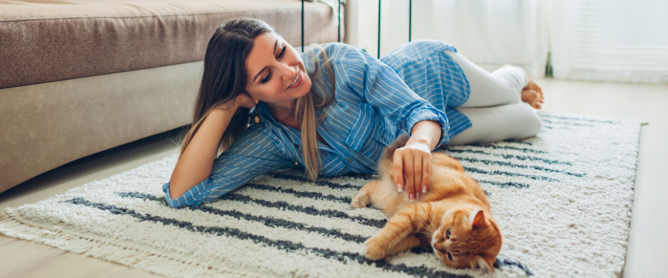 A young woman lying on a rug in a cozy apartment, smiling as she pets an orange cat beside her.