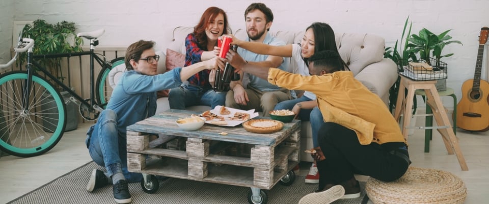 A group of young roommates sitting around a coffee table, enjoying snacks and drinks while toasting together in a cozy apartment living room.