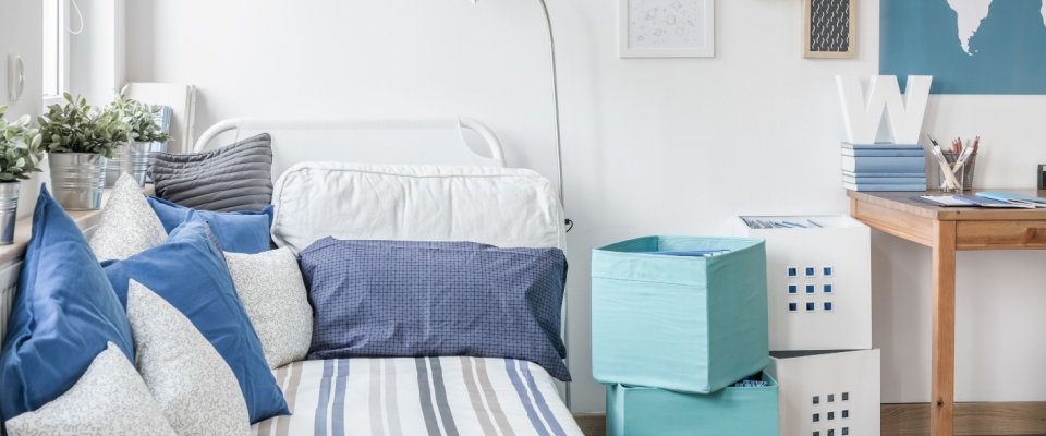 A cozy student bedroom with a bed, pillows, and storage bins.