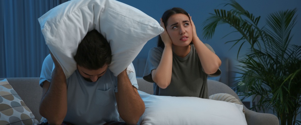 Young couple with pillows suffering from noisy neighbors in living room at night.