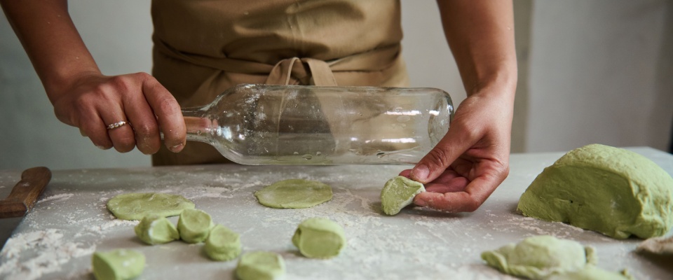 Close-up of a woman in beige apron using an empty glass wine bottle as rolling pin to roll out dough in her kitchen.