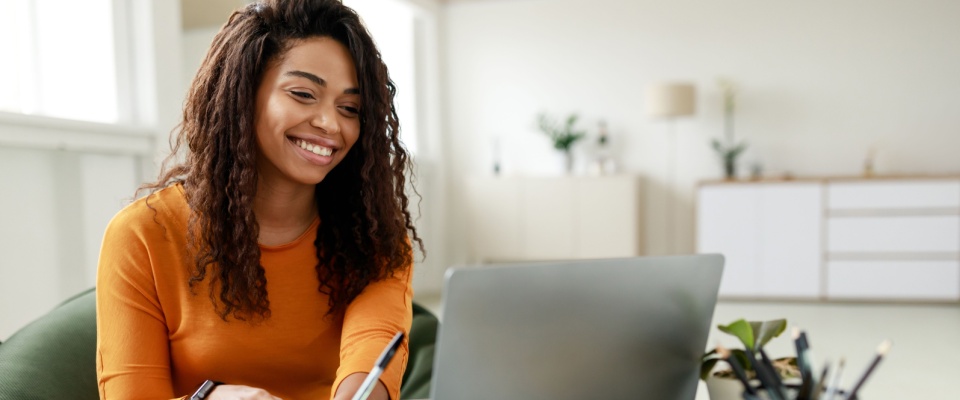 Smiling young African American woman sitting at desk working on laptop taking notes in notebook.
