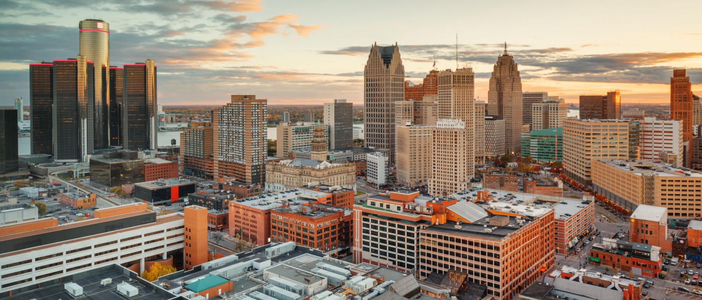 Aerial view of downtown Detroit at sunset, featuring the Renaissance Center and a blend of modern skyscrapers and historic buildings bathed in a warm, golden light.