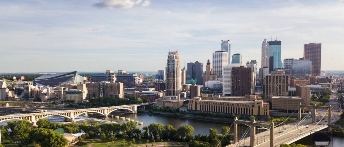 Skyline of downtown Minneapolis, MN, with prominent buildings, bridges, and a river under a clear sky.