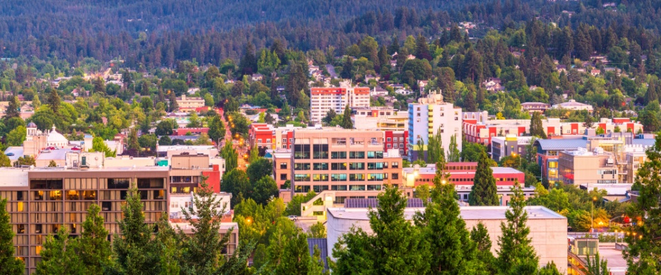 A scenic view of Eugene, Oregon, featuring a mix of modern and historic buildings surrounded by lush green trees and forested hills, highlighting the city's natural beauty and vibrant urban landscape.