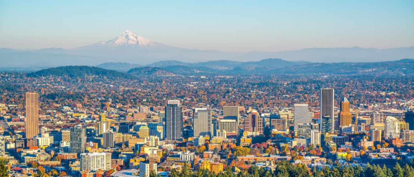 Panoramic view of Portland, Oregon's skyline with Mount Hood visible in the background, framed by lush, forested hills and a mix of modern and historic buildings.