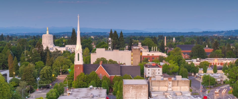 A view of downtown Salem, Oregon, featuring tree-lined streets, the historic Oregon State Capitol building, and a church steeple, with the Cascade Mountains faintly visible in the background.