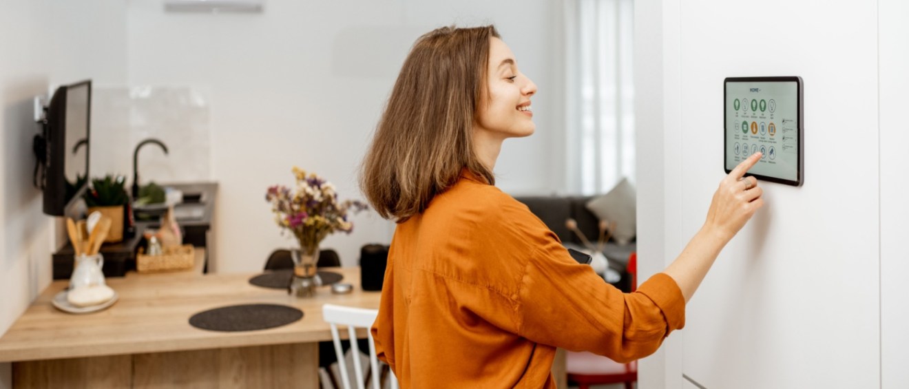 A woman in an orange shirt is smiling as she interacts with a smart home automation panel mounted on the wall in her modern rental apartment.