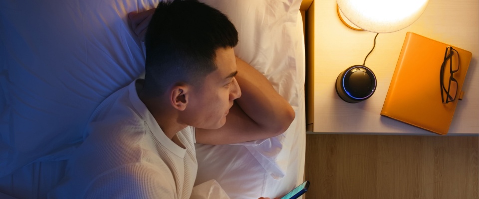 A man lying in bed is speaking to a voice assistant device on his bedside table, using it to control the lights in his home.
