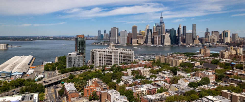 An aerial view of Brooklyn, NY on a beautiful day with blue skies and white clouds. The New York City skyline is in the background featuring the Freedom Tower and lower Manhattan.