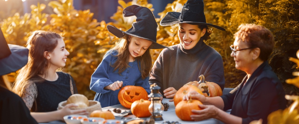 A group of children joined by one adult woman and an older one, gathered around a table and carving some pumpkins for Halloween.