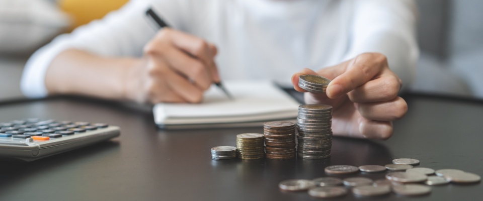 A person writing on a notepad and counting coins, with a calculator within reach.