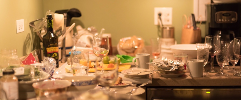 The aftermath of a gathering, where a kitchen counter is covered in large amounts of glasses, plates and silverware.