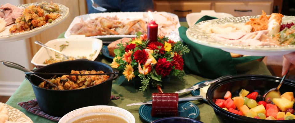 A Thanksgiving-style potluck meal, with plenty of food containers on a table and a candle & flower arrangement in the middle of the table.