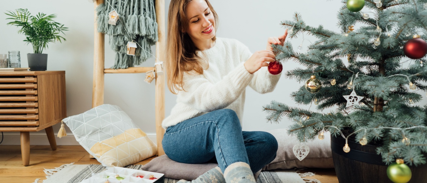 A young woman in a cozy sweater carefully decorating a potted Christmas tree with small glass baubles.