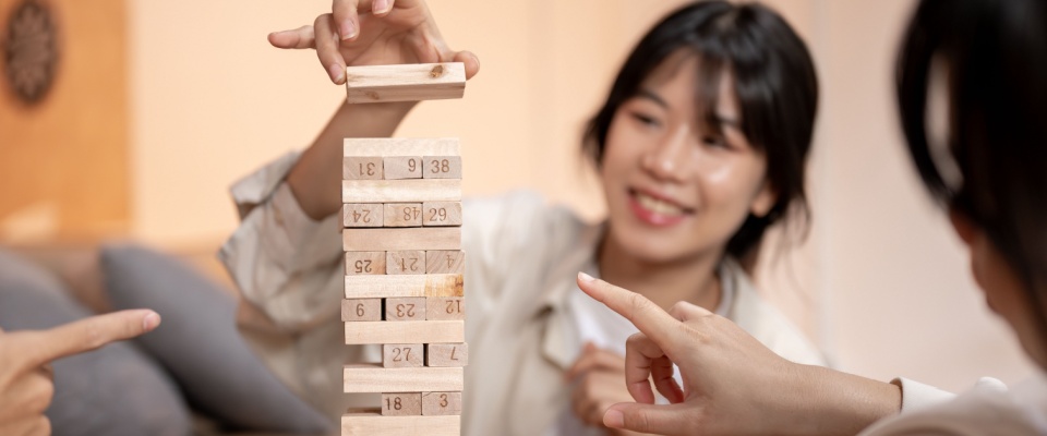 A close-up, selective-focus image of a woman’s hand carefully pulling a wooden block from a Jenga tower while playing with friends at home. 