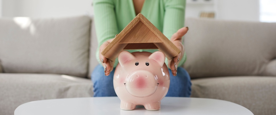 Person holding a small wooden roof over a piggy bank, symbolizing financial savings and protection in rental housing.