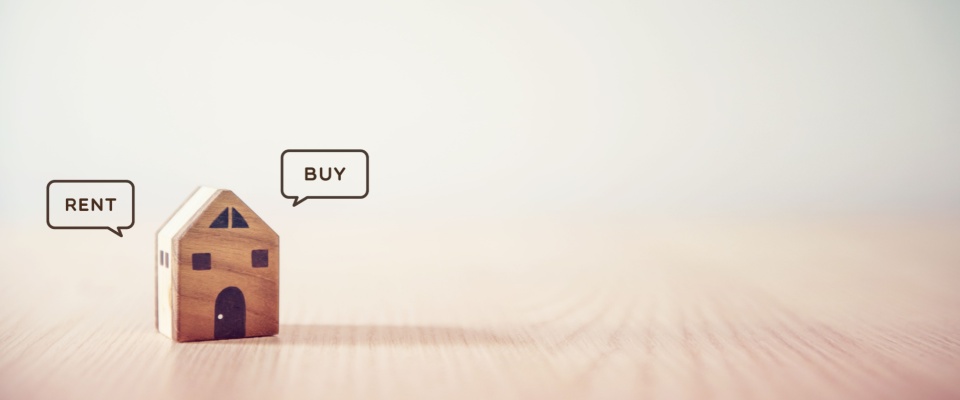 Wooden house model with speech bubbles saying 'Rent' and 'Buy,' representing the choice between renting and buying a home.