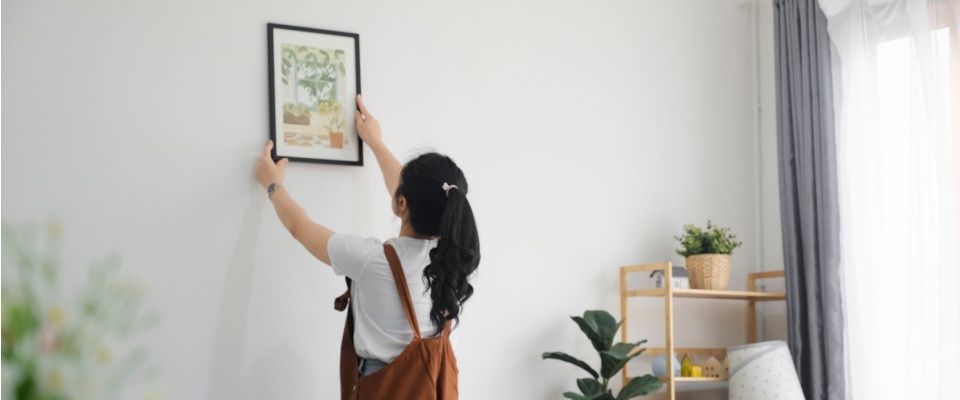 Young woman hanging a framed picture on the wall, adding personal touches to decorate her rental apartment.