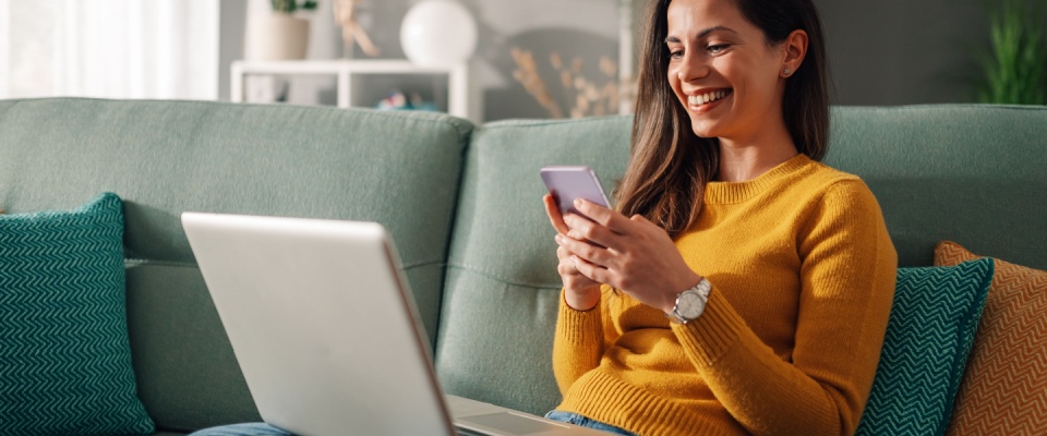 Portrait of a woman using laptop and a phone and checking email or news online while sitting on sofa at home.