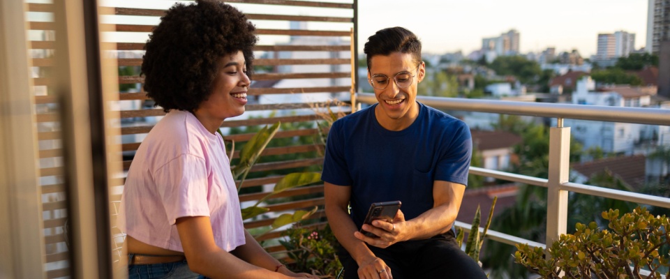Two Latin friends sitting on the balcony looking at the mobile phone and smiling with a view of the city in the background.