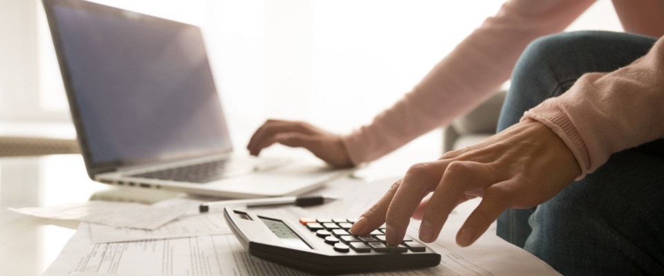 A person uses a laptop and calculator on a low table to keep track and manage expenses.
