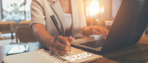 A person sits at their desk, with a laptop in front of them, while also holding a pen in their right hand, above a notebook marked for 2025.