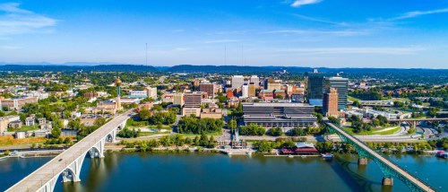 A view of Knoxville, TN, with two of its bridges over the Tennessee River visible in the lower half of the image, and the city’s downtown area seen in the middle.