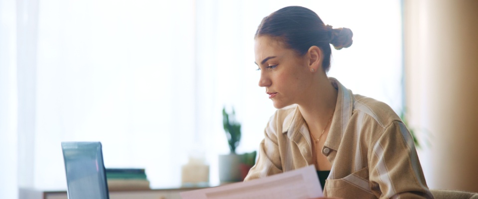A person looking at their laptop with some paperwork in their hand.