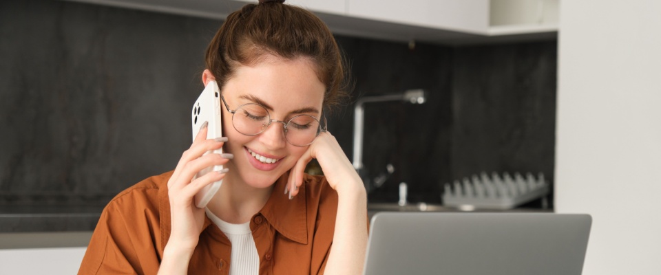 A portrait of a young self-employed woman working from home as a freelancer. She is sitting in her kitchen with a laptop, talking to a client on the phone. The scene captures her professionalism and multitasking as she makes an order or discusses business.
