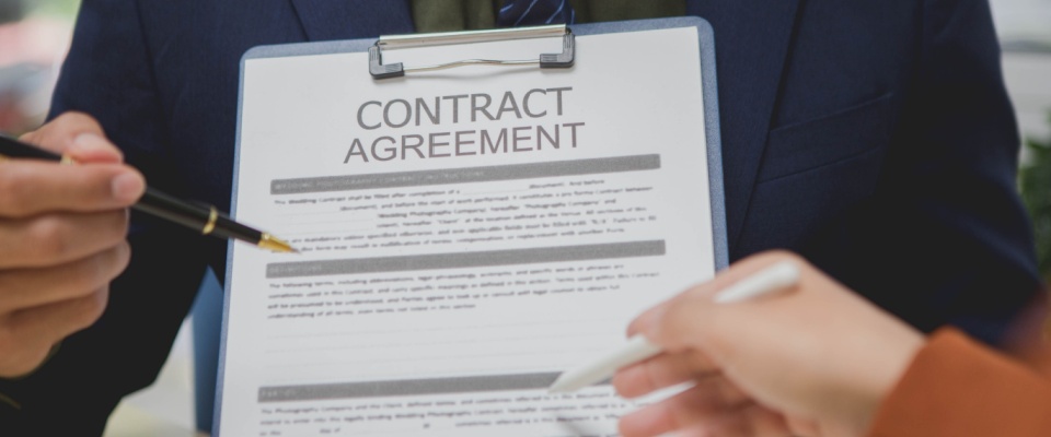 A person holds a clipboard containing a draft of a contract agreement, using the pen in their left hand to point to its contents, while another person, holding a pen in their right hand, goes in closer to sign the paperwork.
