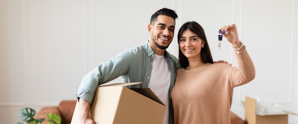 A young couple holding a box and the keys to their new apartment during the process of moving into their new home.